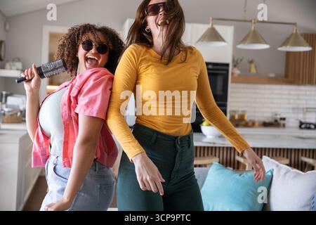 Verschiedene Freundinnen tanzen und singen in der offenen Küche mit Haarbürstenmikrofon und Sonnenbrille Stockfoto Verschiedene Freundinnen tanzen und singen in der offenen Küche mit Haarbürstenmikrofon und Sonnenbrille Stockfoto