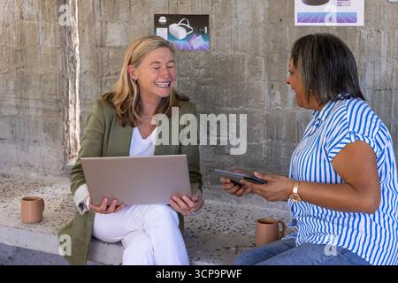 Ältere weibliche Kollegen, die in der Bürolounge unter Technikposter an Laptop und Tablet arbeiten Stockfoto