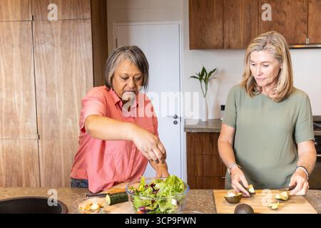 Verschiedene Freundinnen streuen Garnelen und Gurken in eine Schüssel und schneiden Avocado in der Küche Stockfoto