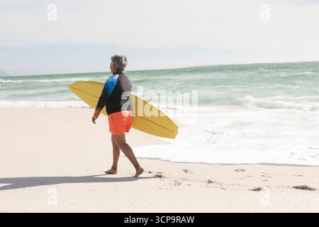 Senior Mann, der barfuß am Sandstrand läuft, trägt blauen Hautschutz und trägt gelbes Surfbrett Stockfoto