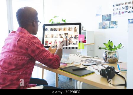 Afroamerikanischer Mann sitzt am kreativen Studiotisch und überprüft Miniaturansichten auf dem Computer mit einem Tablet Stockfoto