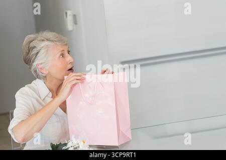 Ältere Frau, die eine rosa Geschenktüte hält, während sie hineinblickt, an einem modernen Küchentisch, Kopierraum Stockfoto