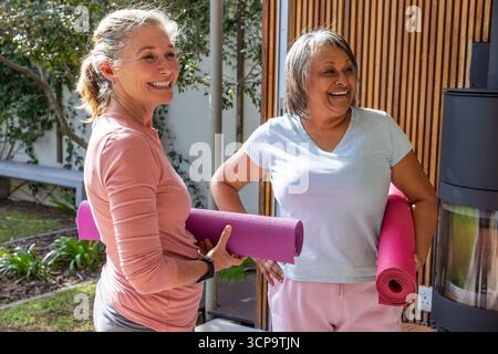 Ältere Freundinnen halten rosafarbene, gerollte Yogamatten und lächeln auf der sonnigen Terrasse neben der Bank Stockfoto