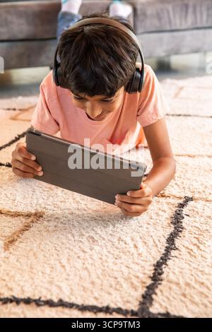 südasiatischer Junge, der auf gemustertem Teppich im Wohnzimmer in schwarzen Kopfhörern mit Tablet liegt Stockfoto