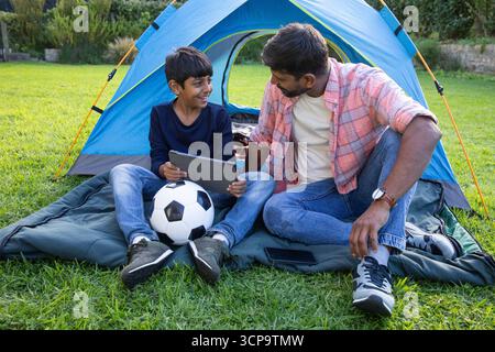 Asiatischer Vater und Sohn sitzen auf Schlafsack vor blauem Zelt mit Tablette und Fußball Stockfoto