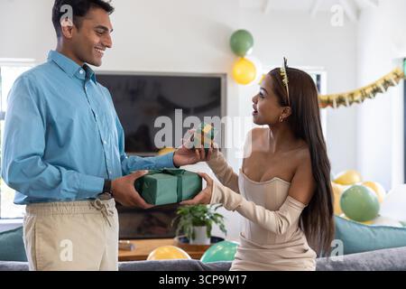 Verschiedene Paare tauschen eingewickelte Geschenke in der Nähe des Sofas im Wohnzimmer mit Ballons und Banner aus Stockfoto