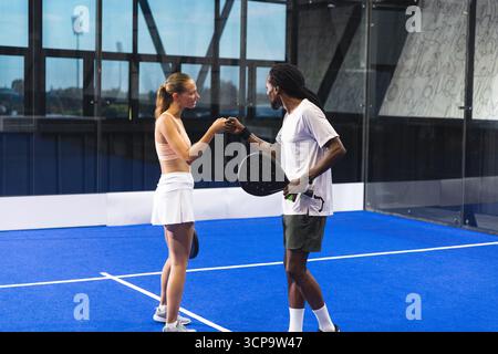 Verschiedene Padel-Partner in Sportbekleidung Faustschläge in der Mitte des blauen Platzes mit Schlägern und Ball Stockfoto