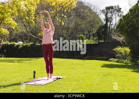 Frau, die Arme in Trainingskleidung auf einer lavendelfarbenen Yogamatte im Garten neben einer schwarzen Wasserflasche streckt Stockfoto