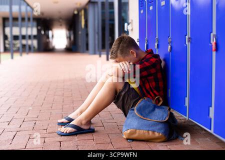 Junge im Schulalter, der auf dem Schulflur sitzt und sich an blaue Schließfächer mit Rucksack und Kopierraum lehnt Stockfoto