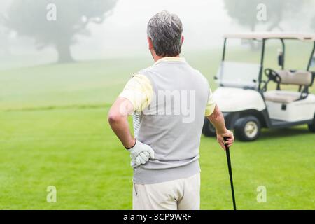 Älterer männlicher Golfspieler, der auf dem Fairway steht und einen Golfschläger trägt, der einen Golfhandschuh in der Nähe eines weißen Golfwagens trägt Stockfoto