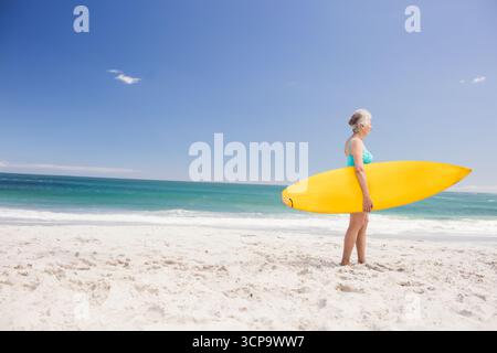 Ältere Frau mit gelbem Surfbrett in blauem Badeanzug am Strand mit Blick auf das türkisfarbene Meer, Kopierraum Stockfoto