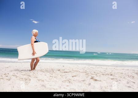 Eine ältere Frau, die barfuß am Strand steht und eine Schaumstoffplatte mit Blick auf das türkisfarbene Meer hält, Kopierraum Stockfoto