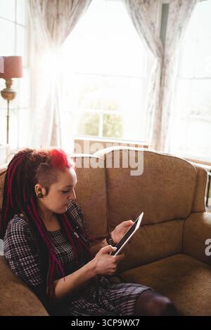 Frau mit bunten Dreadlocks, die Tablette auf beigefarbenem Sofa im sonnendurchfluteten Wohnzimmer hält, Kopierraum Stockfoto