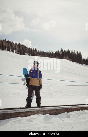 Frau, die auf dem Förderband wartet, trägt Helm-Brille-Pack, das Snowboard an blauer Seilbarriere hält Stockfoto