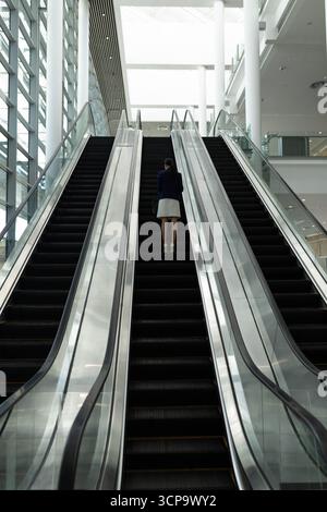 Asiatische Geschäftsfrau, die im modernen Atrium eine Rolltreppe hochfährt und Handtasche an Glasgeländern hält Stockfoto
