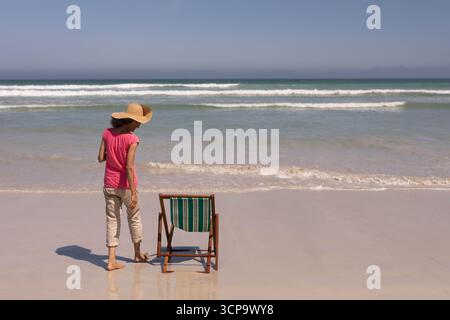Reife Erwachsene Frau, die an der Küste steht und einen Sonnenhut neben einem gestreiften Stuhl trägt, der Schatten reflektiert Stockfoto