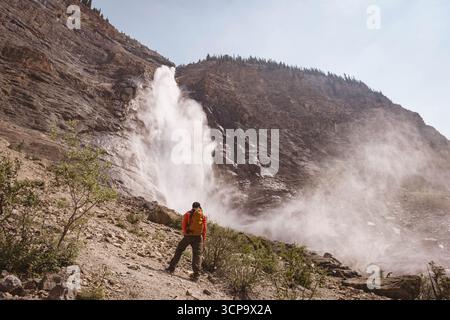 Männlicher Wanderer, der am Hang am Wasserfall steht, in orangefarbener Jacke und mit gelbem Rucksack, Kopierraum Stockfoto