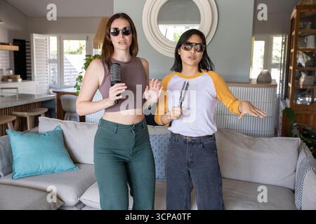 Verschiedene Freundinnen singen in die Haarbürsten, während sie eine Sonnenbrille in einem offenen Wohnzimmer tragen Stockfoto Verschiedene Freundinnen singen in die Haarbürsten, während sie eine Sonnenbrille in einem offenen Wohnzimmer tragen Stockfoto