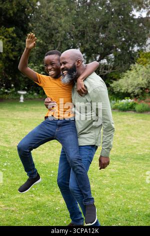 afroamerikanischer Vater und Sohn spielen im Garten, tragen den Jungen auf dem Rücken, winken in der Nähe des Vogelbades Stockfoto