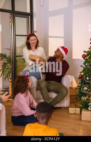 Verschiedene Familien tauschen Geschenke zu Hause in der Nähe des weihnachtsbaums aus, tragen weihnachtsmütze, Geweih-Haarbänder Stockfoto