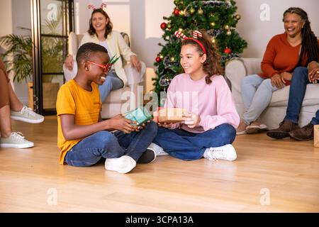 Verschiedene Kinder, die im Wohnzimmer unter dem weihnachtsbaum in der Nähe von Sofas Geschenke austauschen Stockfoto