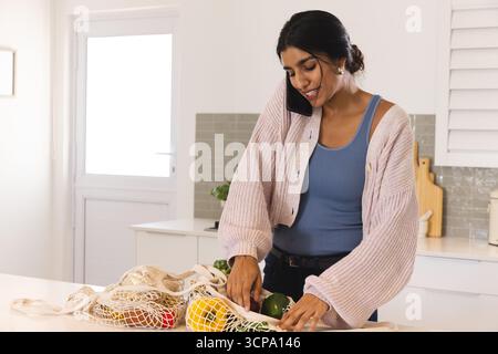 Erwachsene südasiatische Frau packt auf der Kücheninsel eine Tasche mit Zitronen und Paprika aus und hält ein Smartphone Stockfoto