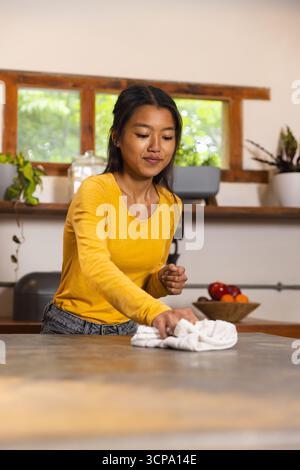 Asiatische Frau, die Küchenarbeitsplatte mit weißem Tuch in der Nähe einer hölzernen Obstschale in der Wohnküche reinigt Stockfoto