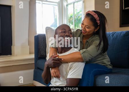 Diverse Paare umarmen und lächeln auf blauem Sofa im Wohnzimmer neben dem großen Fenster Stockfoto