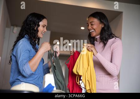 Verschiedene Freundinnen halten und vergleichen grünes Tanktop, rotes Hemd, gelbe Bluse im Flur Stockfoto