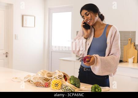 Südasiatische Frau, die auf dem Smartphone spricht, roten Pfirsich in der Küche hält, mit Netztasche, Kopierraum Stockfoto
