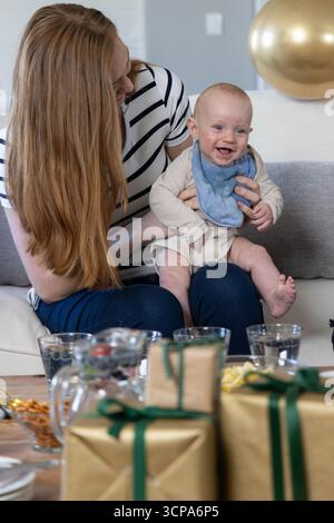 Mutter und Baby sitzen auf der Heimcouch und schmiegen lächelndes Kind neben goldenen Geschenken, Ballon Stockfoto