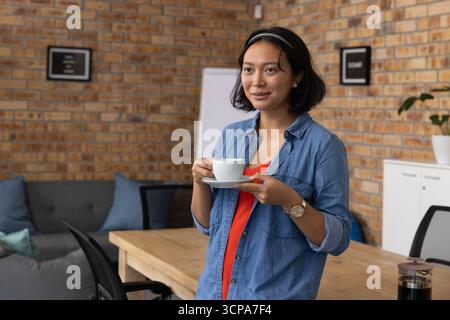 Asiatische Frau mit weißer Kaffeetasse neben Tisch mit französischer Presse im Büro, Kopierraum Stockfoto