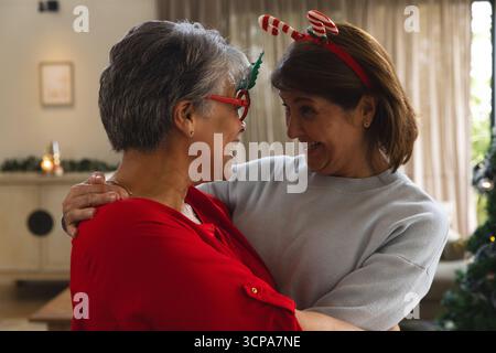 Vielfältige Mutter und Tochter umarmen sich zu Hause am Weihnachtsbaum mit Geweih und Zuckerrohr Stirnbänder Stockfoto