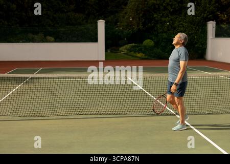 Senior Mann, der auf dem Tennisplatz steht und eine Schläger-Sportuhr hält und nach oben blickt, Kopierraum Stockfoto