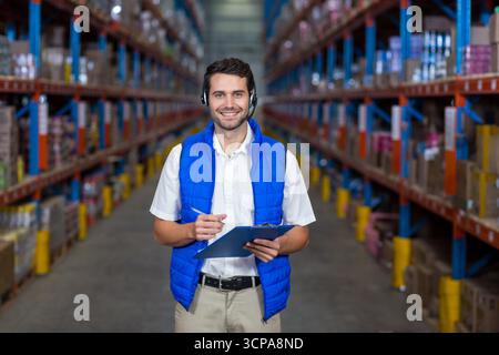 Männlicher Lagerarbeiter im Gang, flankiert von Regalen mit gestapelten Boxen, mit Klemmbrett Stockfoto