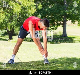 Mann, der auf dem Rasen im Park steht und sich zu den Knöcheln dehnt, trägt ein rotes ärmelloses Oberteil mit Laufschuhen Stockfoto