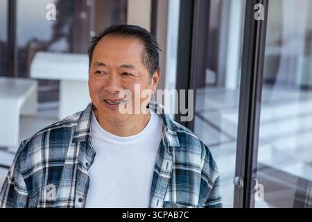 Asiatischer Seniorenmann lächelte, während er auf der Terrasse neben Bank und Glasschiebetüren blickte Stockfoto