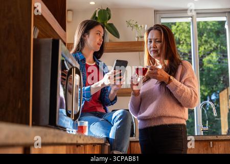 Asiatische Mutter und Tochter plaudern auf der Kücheninsel mit Smartphone und einer Tasse Tee Stockfoto