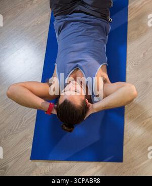 Mann, der auf der Matte liegt und auf dem Boden des Fitnessraums knirscht, mit ärmellosem Top-Fitness-Tracker Stockfoto
