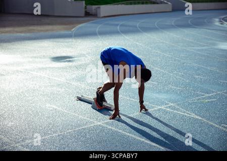 Afroamerikanischer erwachsener männlicher Athlet, der sich im Sprint auf der blauen Strecke mit Metallblöcken ausrichtet Stockfoto