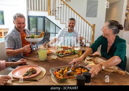 Verschiedene Freunde sitzen zu Hause um den Tisch und teilen sich Gemüse und Salatschüssel Stockfoto