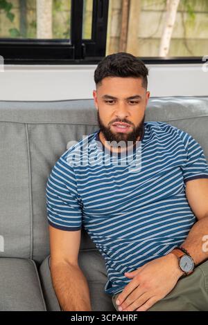 Bärtiger Mann sitzt auf Sofa unter schwarz gerahmtem Fenster im Wohnzimmer und trägt Armbanduhr Stockfoto