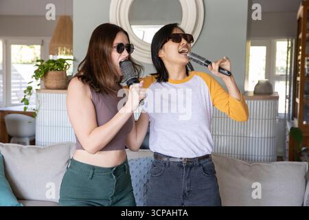 Verschiedene Freundinnen mit Haarbürsten, die Karaoke im Wohnzimmer mit Spiegel und Pflanze vortäuschen Stockfoto Verschiedene Freundinnen mit Haarbürsten, die Karaoke im Wohnzimmer mit Spiegel und Pflanze vortäuschen Stockfoto