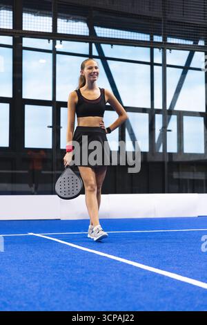 Weibliche Athletin, die auf dem blauen Padel-Tennisplatz steht und einen Padel-Schläger hält, lächelt mit Glaswänden Stockfoto
