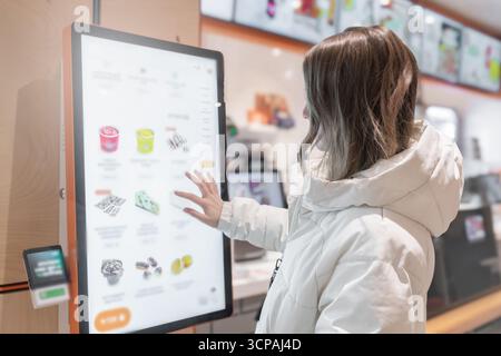 Eine Frau in einer weißen Winterjacke bestellt Kuchen und Speisen über einen Touchscreen in einem Fast-Food-Restaurant oder einem Pizzeria-Café. Eine Frau interagiert Stockfoto