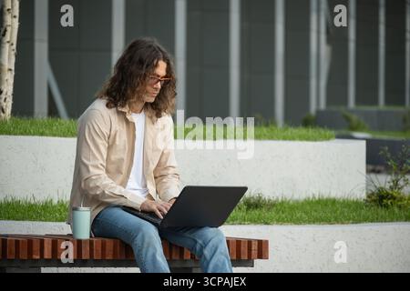 Junger Mann, Student, attraktiver Freiberufler sitzt auf einer Bank im Stadtpark und nutzt Laptop für Fernarbeit und Bildung. Im Freien. Stockfoto