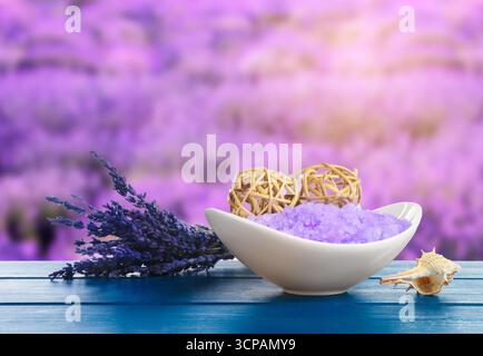 Meersalz in der Schüssel und Blumen auf blauem Holztisch vor verschwommenem Lavendelfeld Stockfoto