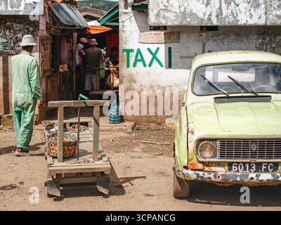 Ambositra, Madagaskar - 26. April 2019: Altes rostiges Auto parkt an einem sonnigen Tag neben der Hauptstraße, TEXTTAXI dahinter, Gruppe unbekannter Einheimischer im Hintergrund Stockfoto