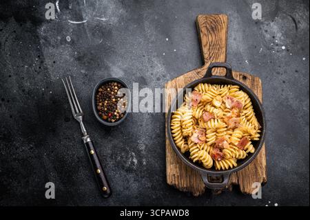 Fusilli Carbonara Nudeln mit knusprigen Speckstückchen und Parmesankäse in einer Pfanne. Schwarzer Hintergrund. Draufsicht. Stockfoto