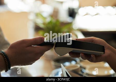 Kaukasischer junger Erwachsener, der ein Smartphone hält und kontaktlos bezahlt, indem er ein drahtloses Terminal benutzt, das von einem anderen gehalten wird, Nahaufnahme der Hände, die digitale Transaktionen im Café durchführen Stockfoto